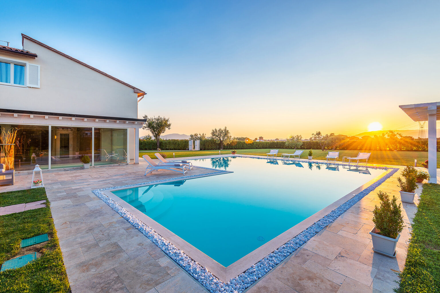 Modern villa with a sparkling swimming pool at sunset, surrounded by lush greenery and comfortable lounge chairs, creating a serene outdoor living space. | Only on SopranoVillas