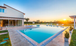 Modern villa with a sparkling swimming pool at sunset, surrounded by lush greenery and comfortable lounge chairs, creating a serene outdoor living space.