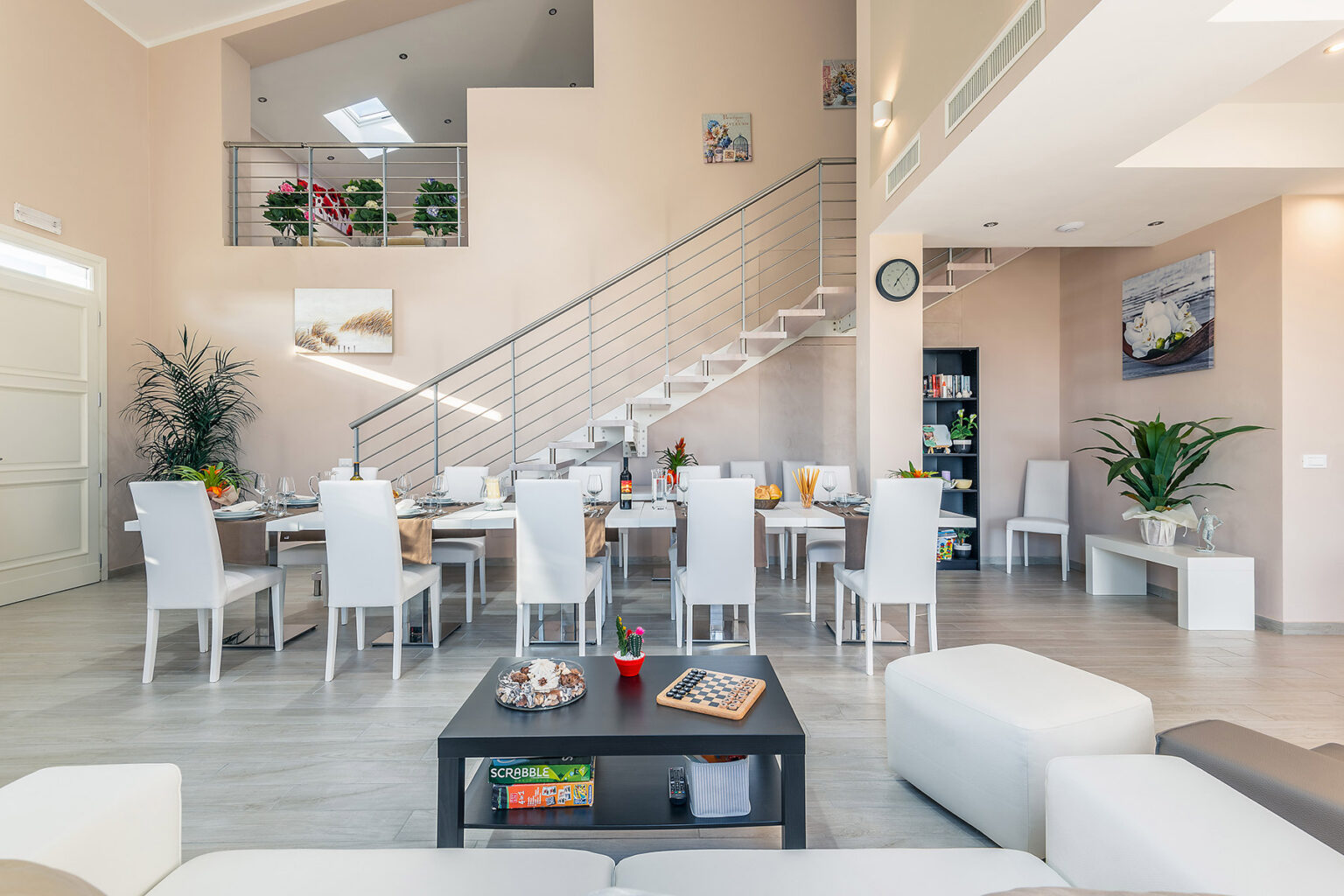 Modern dining area with a large table set for a meal, surrounded by white chairs, and a stylish living room featuring a coffee table and decorative items. The space is bright and airy, highlighted by natural light and contemporary decor. | Only on SopranoVillas