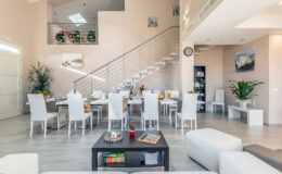 Modern dining area with a large table set for a meal, surrounded by white chairs, and a stylish living room featuring a coffee table and decorative items. The space is bright and airy, highlighted by natural light and contemporary decor.