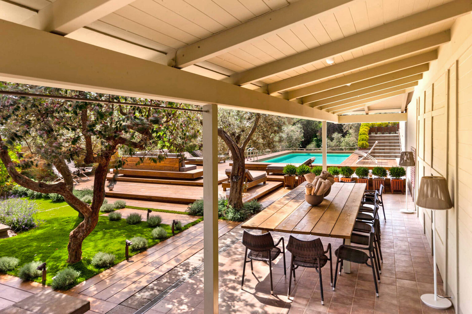 Spacious outdoor dining area with a long wooden table and black chairs, surrounded by lush greenery and trees, overlooking a swimming pool and landscaped garden. | Only on SopranoVillas