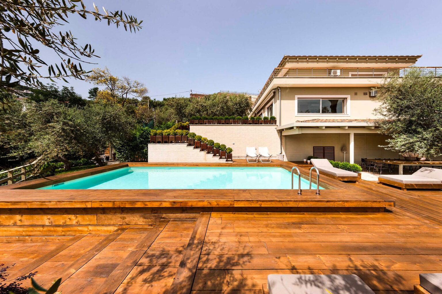 Modern villa with a wooden deck surrounding a sparkling swimming pool, set in a lush garden. The property features a spacious terrace and is surrounded by greenery, creating a serene outdoor living space. | Only on SopranoVillas