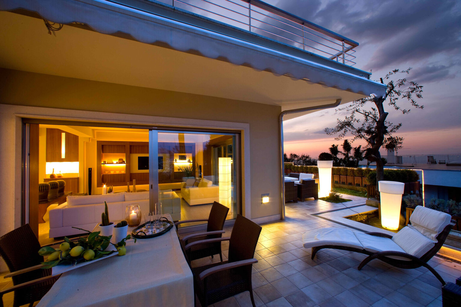 Modern outdoor living space at sunset, featuring stylish lounge chairs, a dining table with fresh fruit, and illuminated planters, showcasing a contemporary home design. | Only on SopranoVillas
