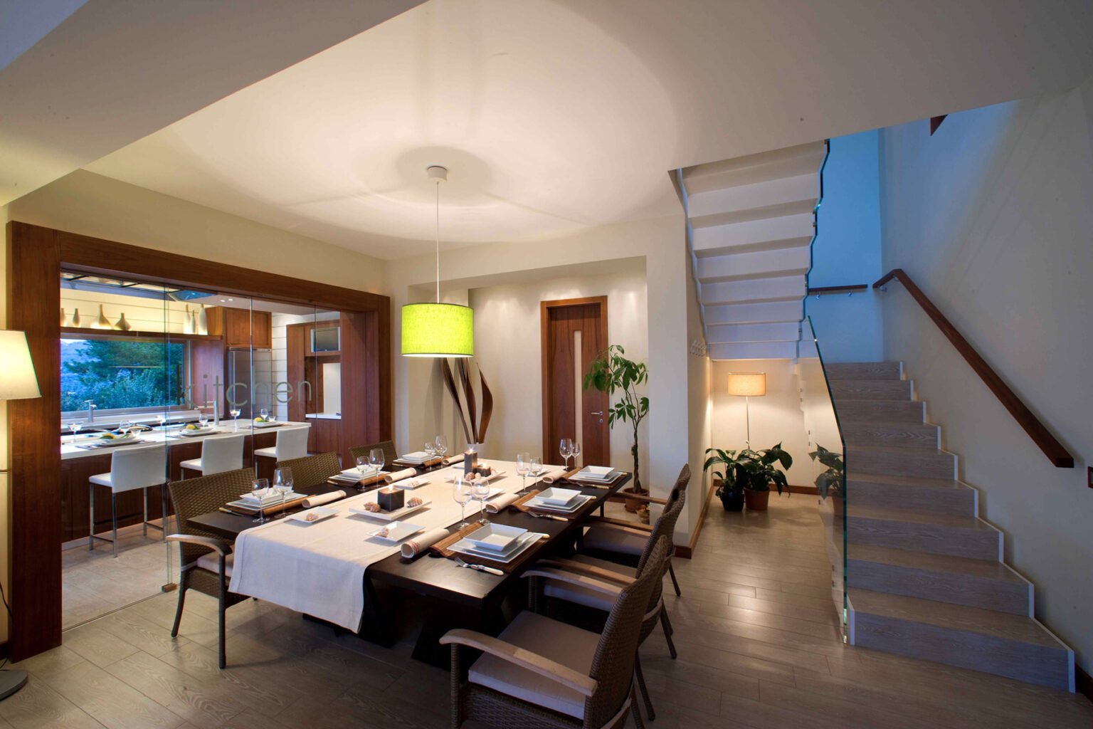 Modern dining room with a large wooden table set for a meal, stylish chairs, and elegant lighting. The open layout connects to a well-equipped kitchen and features a staircase leading to the upper level. | Only on SopranoVillas