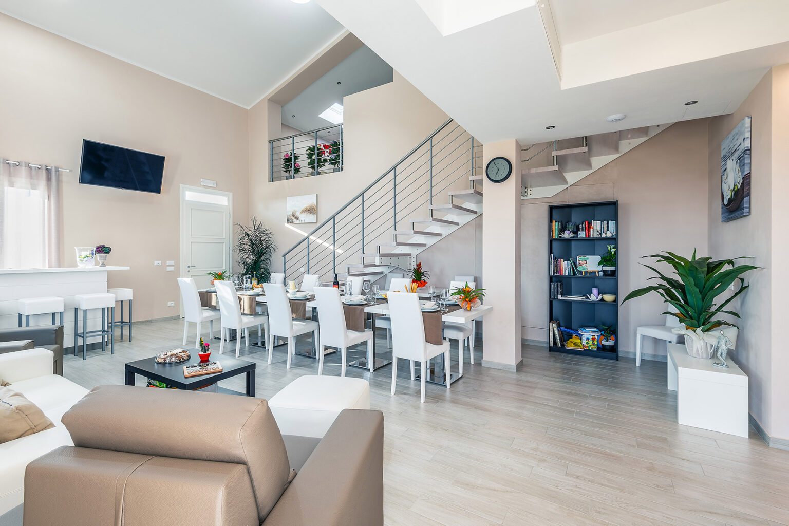 Modern open-concept living area featuring a stylish lounge with a beige sofa, a dining space with white chairs, and a staircase leading to an upper level. The room is bright and airy, adorned with plants and decorative elements, creating a welcoming atmosphere. | Only on SopranoVillas