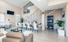 Modern open-concept living area featuring a stylish lounge with a beige sofa, a dining space with white chairs, and a staircase leading to an upper level. The room is bright and airy, adorned with plants and decorative elements, creating a welcoming atmosphere.