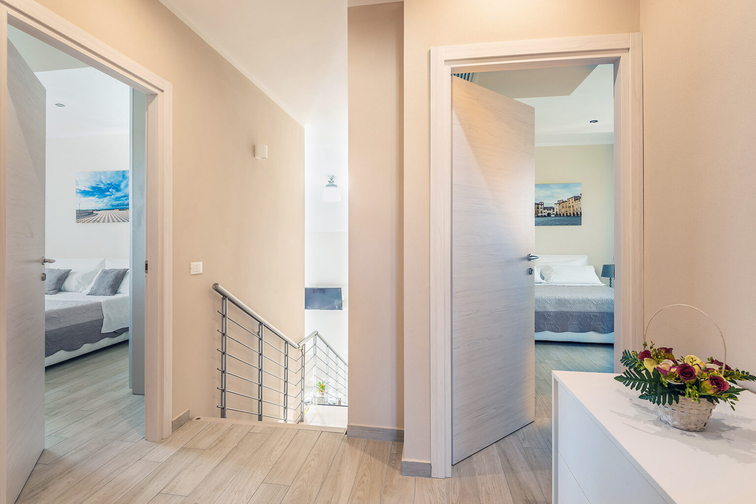 Modern hallway with light-colored walls, featuring open doors leading to bedrooms, a staircase with railing, and a decorative flower arrangement on a console table. | Only on SopranoVillas
