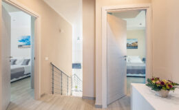 Modern hallway with light-colored walls, featuring open doors leading to bedrooms, a staircase with railing, and a decorative flower arrangement on a console table.