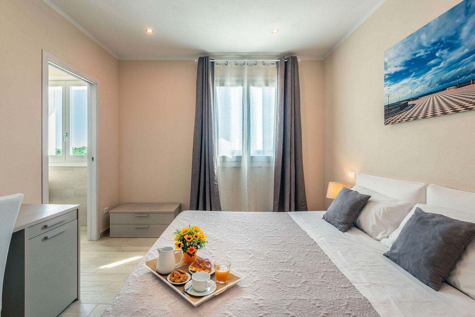 Cozy hotel room featuring a comfortable bed with gray pillows, a breakfast tray with coffee and pastries, and a window with natural light. The decor includes a flower vase and a landscape painting, creating a warm and inviting atmosphere. | Only on SopranoVillas