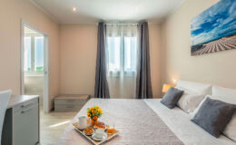 Cozy hotel room featuring a comfortable bed with gray pillows, a breakfast tray with coffee and pastries, and a window with natural light. The decor includes a flower vase and a landscape painting, creating a warm and inviting atmosphere.