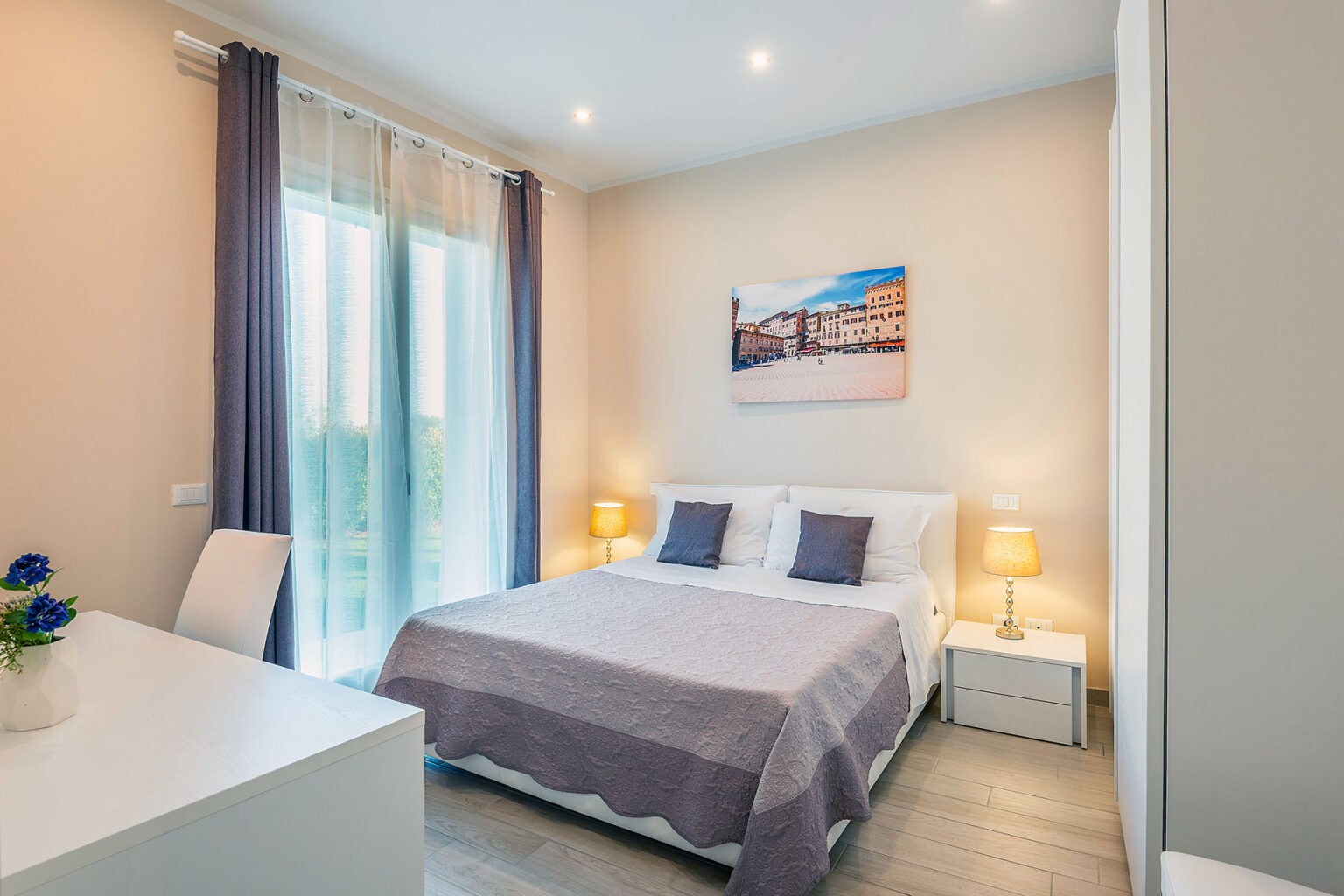 Cozy modern bedroom featuring a comfortable double bed with gray bedding, two bedside lamps, and large windows allowing natural light. A stylish desk and chair complement the space, along with a decorative painting on the wall. Ideal for relaxation and comfort. | Only on SopranoVillas