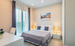 Cozy modern bedroom featuring a comfortable double bed with gray bedding, two bedside lamps, and large windows allowing natural light. A stylish desk and chair complement the space, along with a decorative painting on the wall. Ideal for relaxation and comfort.