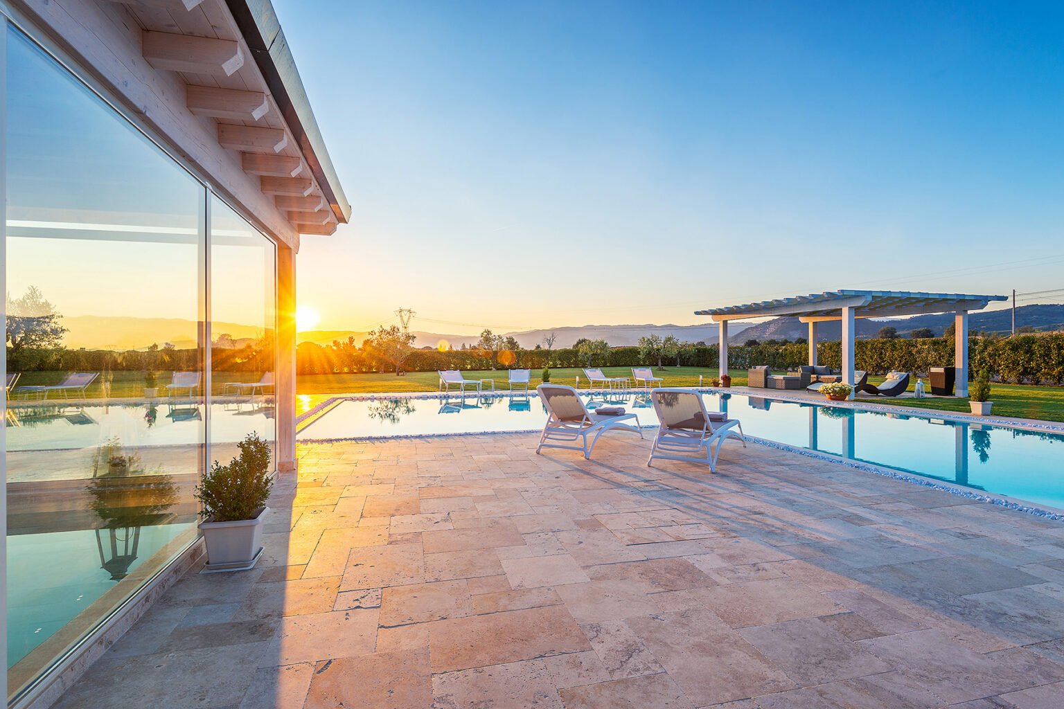 Modern luxury villa with a stunning swimming pool at sunset, featuring lounge chairs and a serene landscape in the background. | Only on SopranoVillas