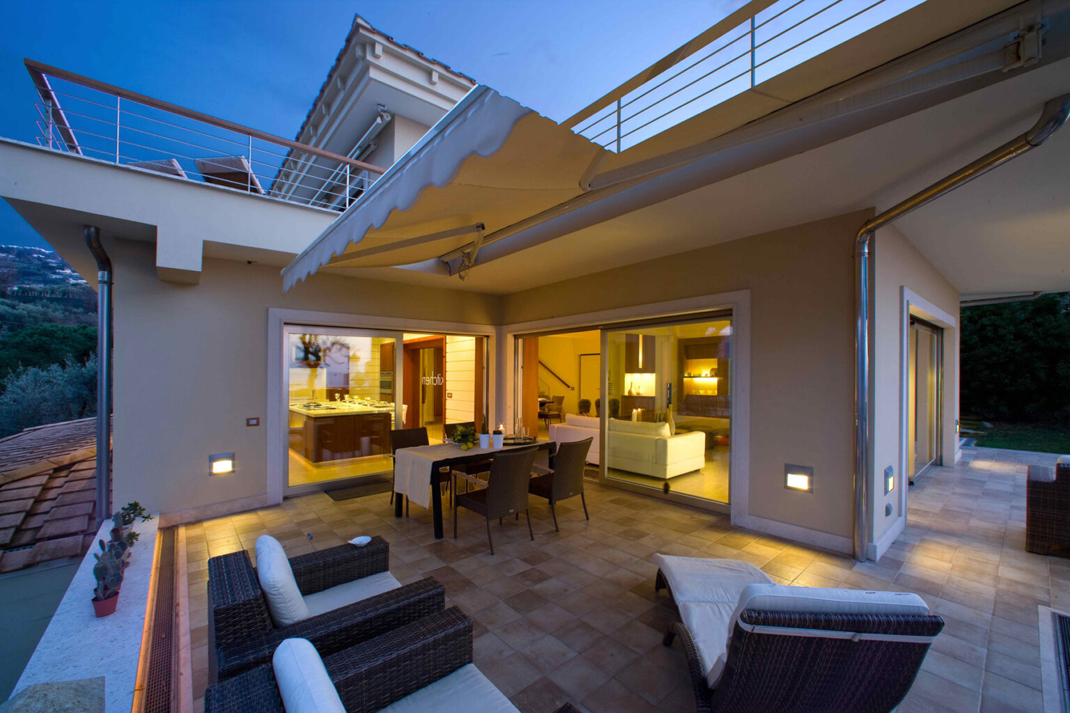 Modern outdoor living space featuring a stylish patio with seating, dining area, and large glass doors leading into a contemporary interior, illuminated at dusk. | Only on SopranoVillas