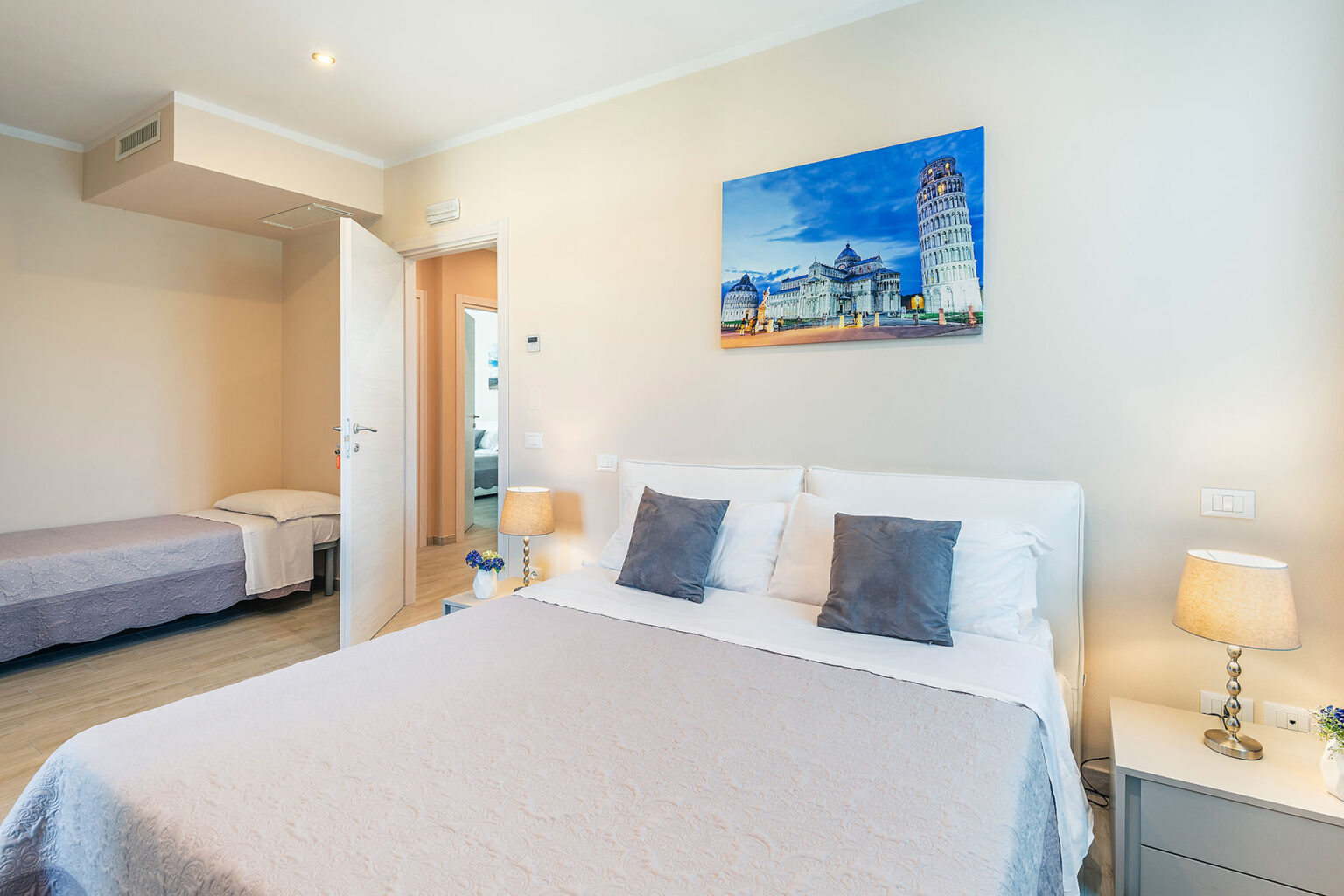 Cozy and modern hotel room featuring a queen-sized bed with gray accent pillows, a twin bed in the background, bedside lamps, and a colorful wall art depicting a scenic view. The room is well-lit with natural light and has a clean, minimalistic design. | Only on SopranoVillas