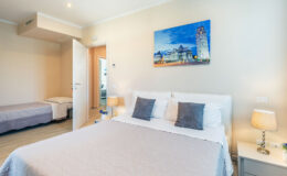 Cozy and modern hotel room featuring a queen-sized bed with gray accent pillows, a twin bed in the background, bedside lamps, and a colorful wall art depicting a scenic view. The room is well-lit with natural light and has a clean, minimalistic design.
