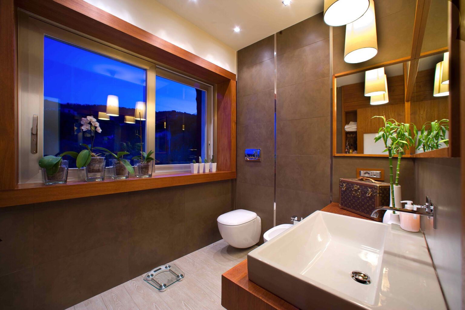 Modern bathroom interior featuring a large window with evening views, elegant wooden accents, a sleek sink, a stylish toilet, and decorative plants, illuminated by warm lighting fixtures. | Only on SopranoVillas