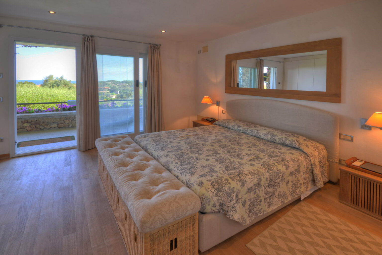 Spacious and modern bedroom featuring a large bed with floral bedding, a stylish mirror, and sliding glass doors that open to a scenic view. The room is well-lit with natural light and includes cozy bedside lamps and wooden furniture. | Only on SopranoVillas
