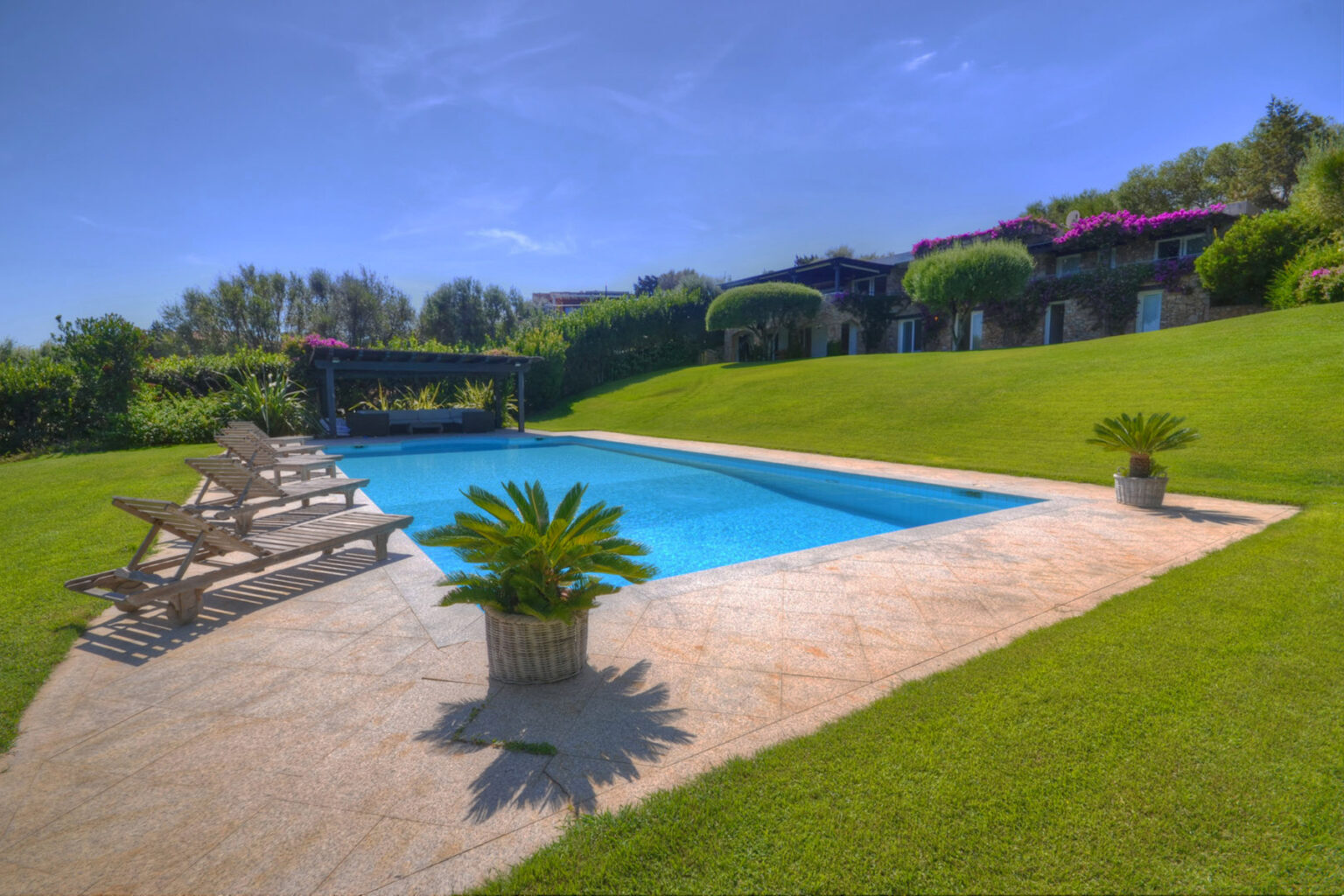 Luxurious outdoor swimming pool surrounded by lush green grass and tropical plants, featuring comfortable lounge chairs and a shaded area, ideal for relaxation and leisure. | Only on SopranoVillas