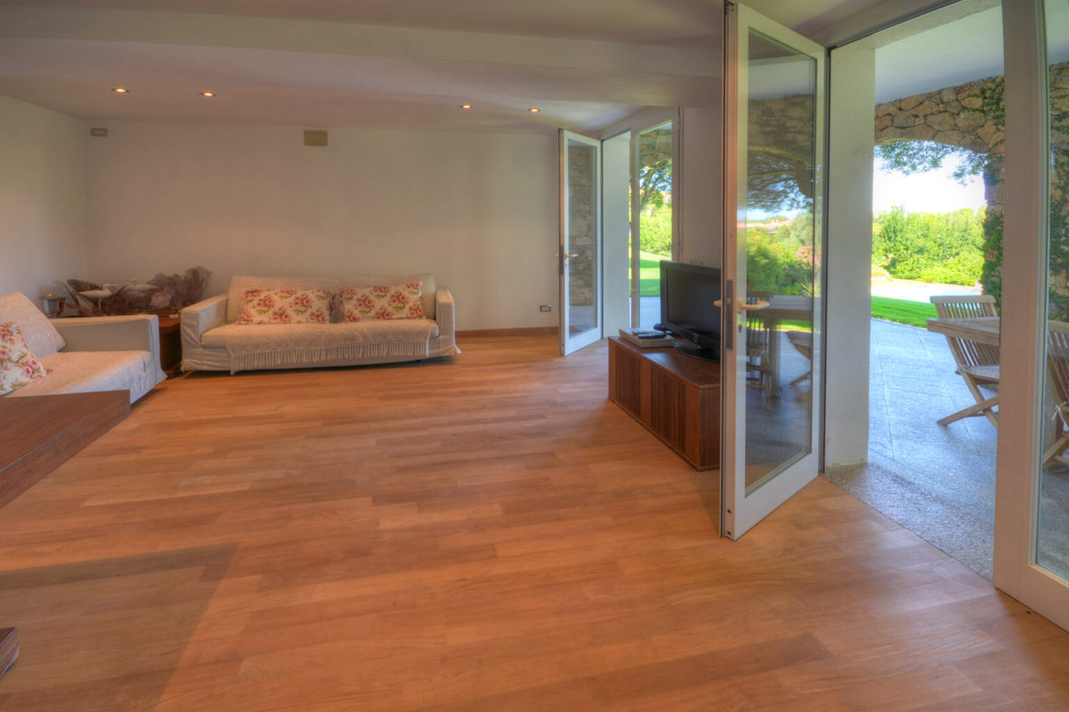 Bright and airy living room featuring hardwood flooring, comfortable sofas with decorative pillows, and large glass doors leading to a lush outdoor area. | Only on SopranoVillas