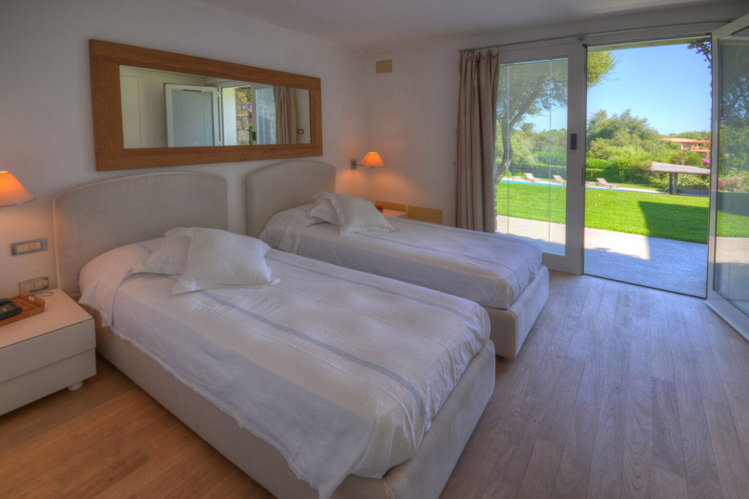 Cozy twin bedroom with modern decor, featuring two beds, bedside table, and large glass doors leading to a lush outdoor area with a pool view. | Only on SopranoVillas