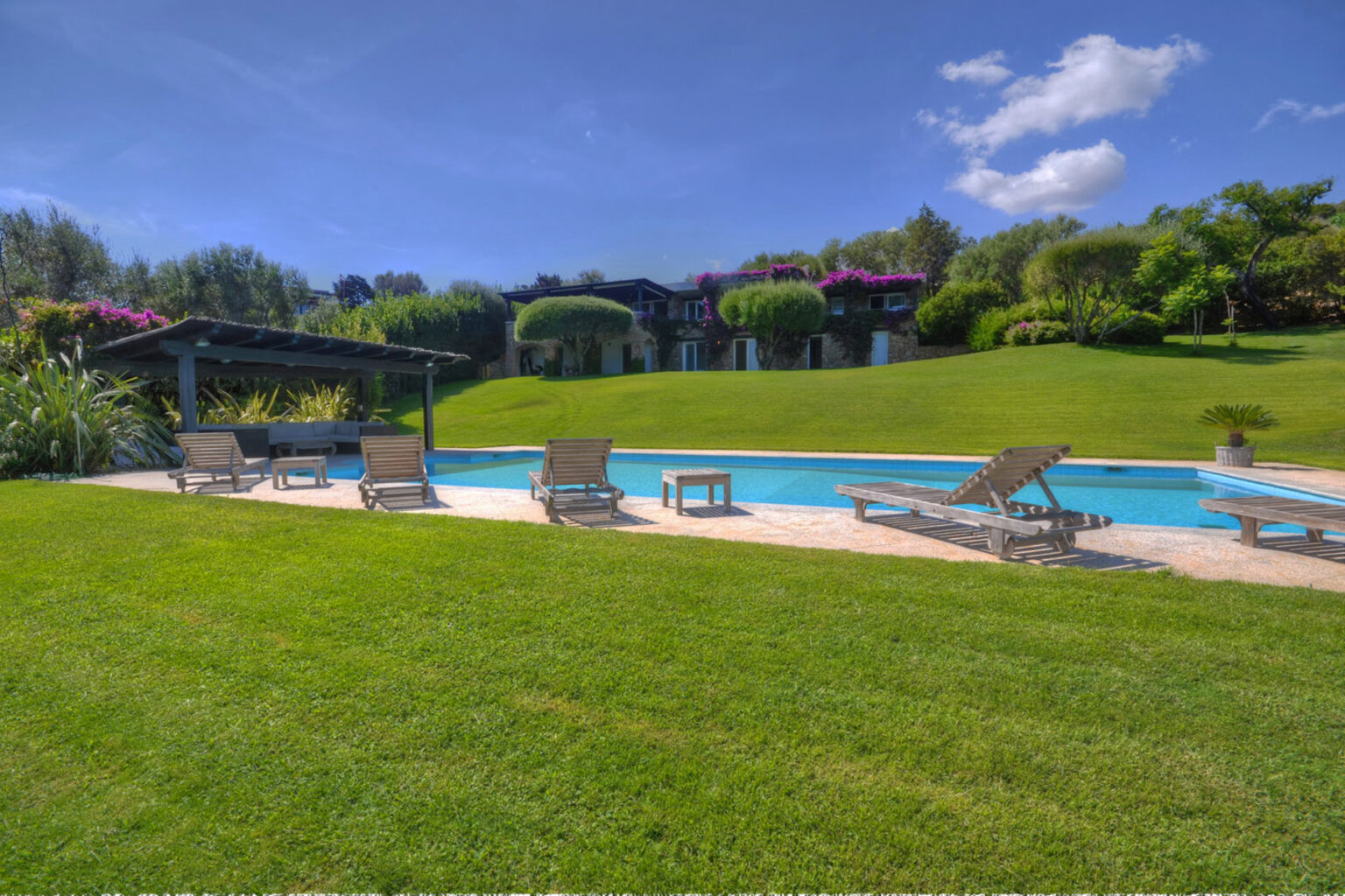 Scenic outdoor swimming pool surrounded by lush green grass and tropical landscaping, featuring lounge chairs and a shaded pergola under a clear blue sky. | Only on SopranoVillas