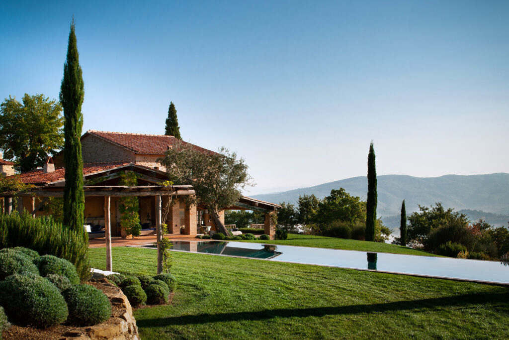 Luxurious villa with a stunning infinity pool overlooking scenic hills, surrounded by manicured gardens and cypress trees under a clear blue sky. | Only on SopranoVillas