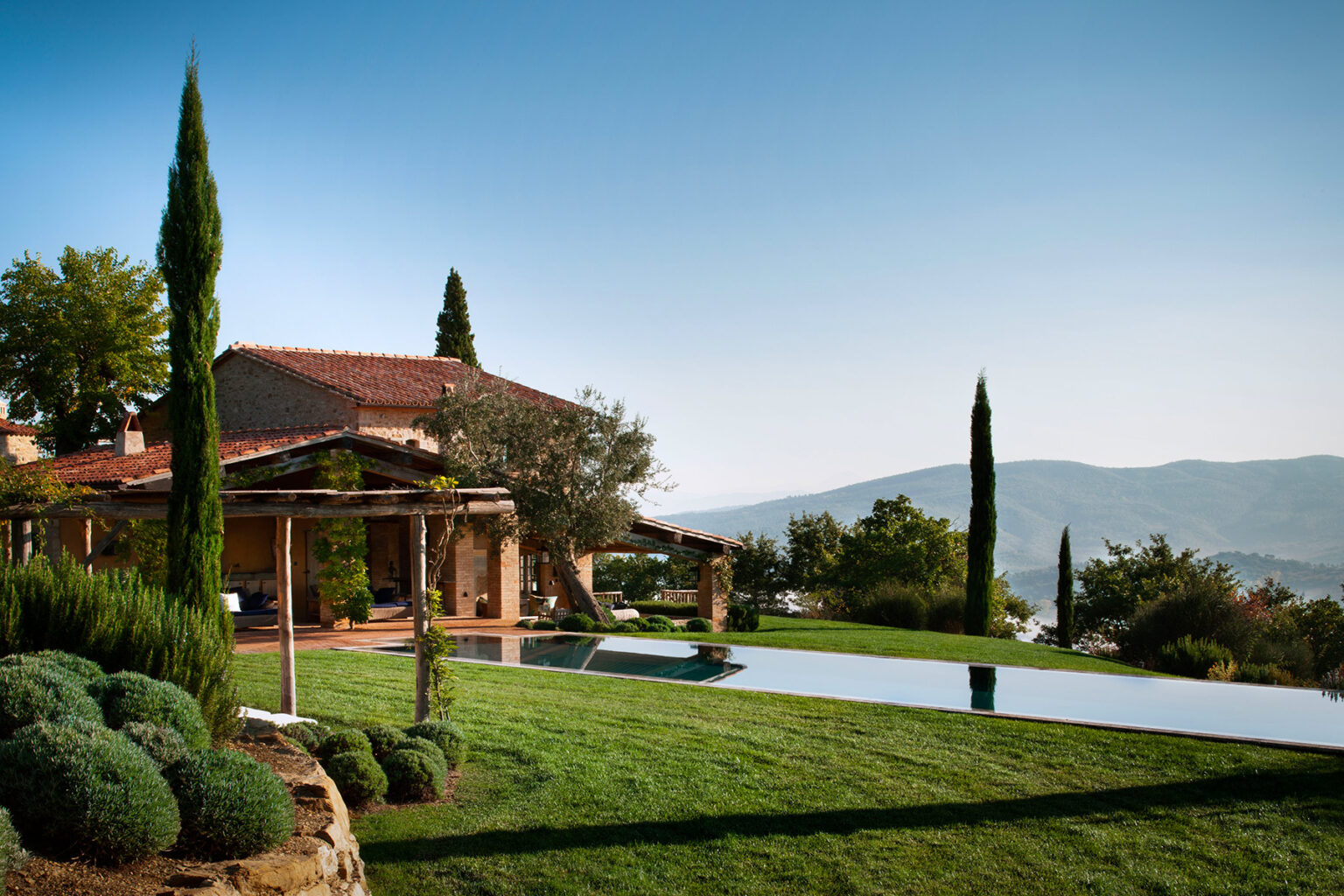 Luxurious villa with a stunning infinity pool overlooking scenic hills, surrounded by manicured gardens and cypress trees under a clear blue sky. | Only on SopranoVillas