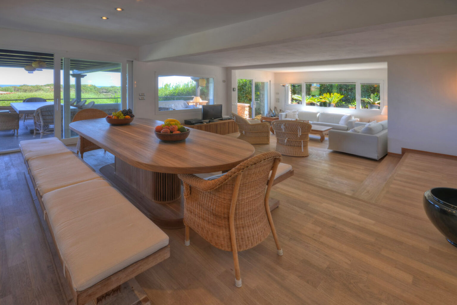 Modern open-concept living room and dining area featuring a large wooden table, wicker chairs, and a bright, airy atmosphere with panoramic views of nature. | Only on SopranoVillas