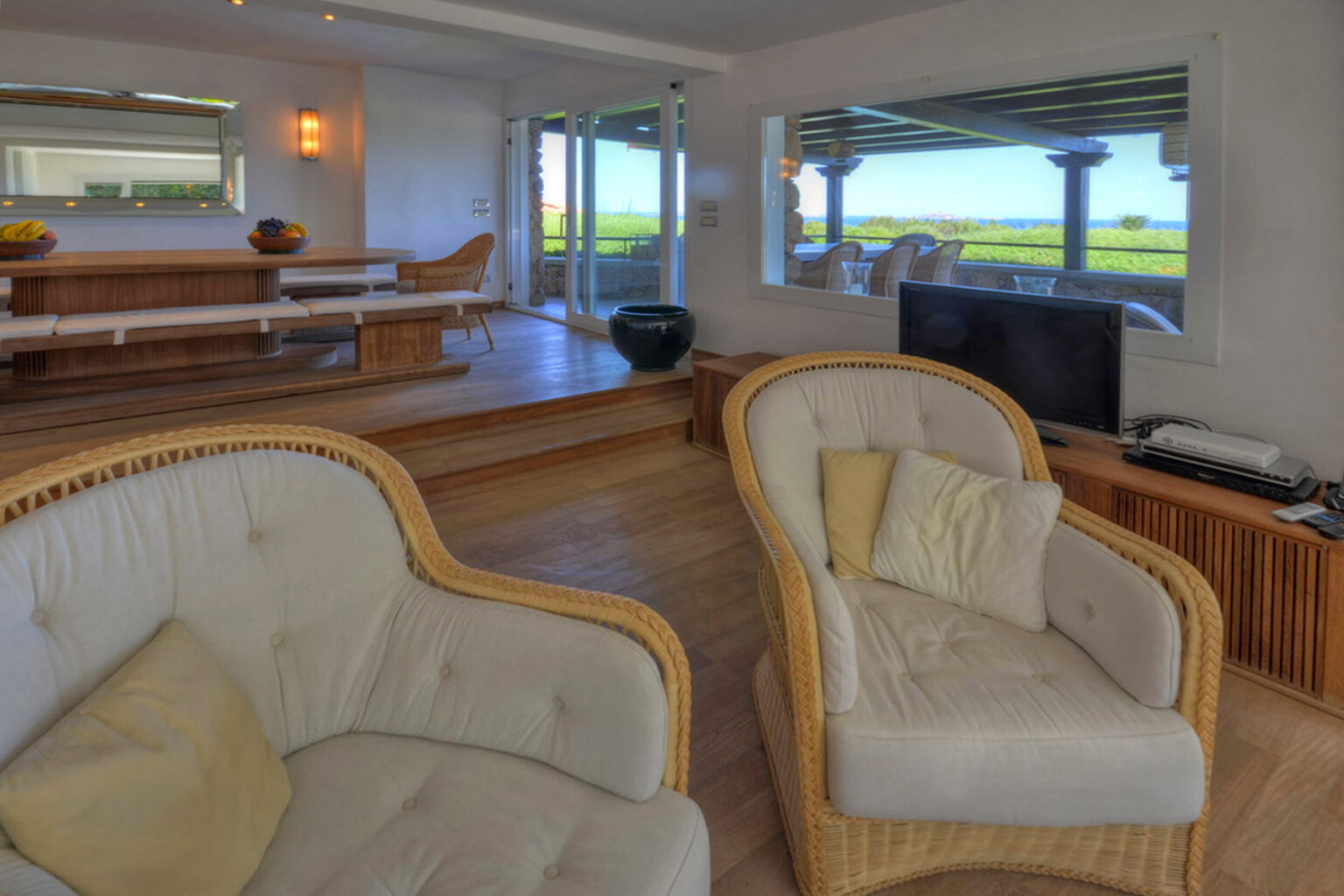 Modern living room interior featuring two comfortable wicker chairs with cushions, a wooden dining area in the background, and large windows showcasing a scenic view of greenery. | Only on SopranoVillas