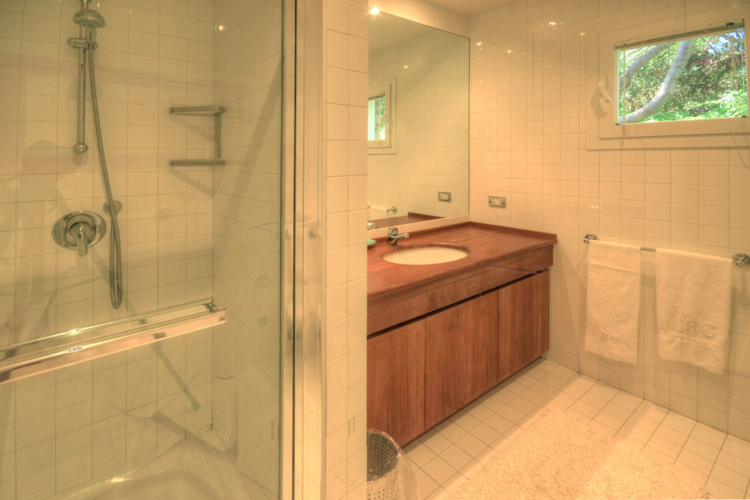 Modern bathroom interior featuring a glass shower enclosure, wooden vanity with a sink, and natural light coming through a window. White tiles and towels add a clean, minimalist aesthetic. | Only on SopranoVillas