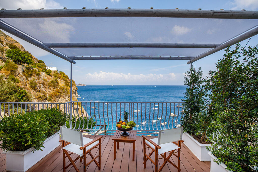 Scenic balcony with two chairs and a table overlooking the ocean, framed by lush greenery and a sunshade, offering a tranquil coastal retreat. | Only on SopranoVillas