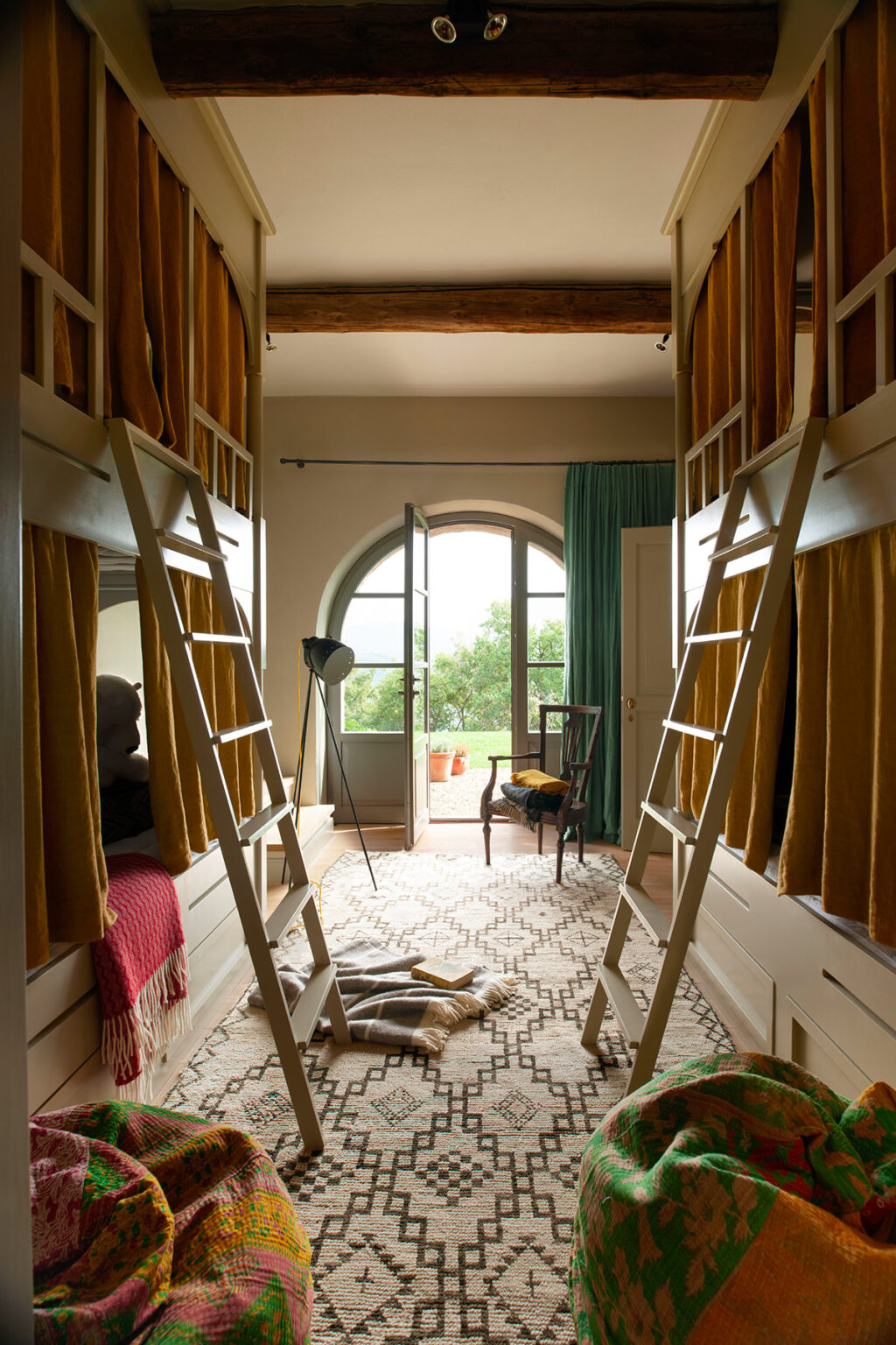 Cozy bedroom interior featuring bunk beds with yellow curtains, a vintage chair, and large windows offering a view of greenery outside. The room is decorated with a patterned rug and a standing lamp, creating a warm and inviting atmosphere. | Only on SopranoVillas