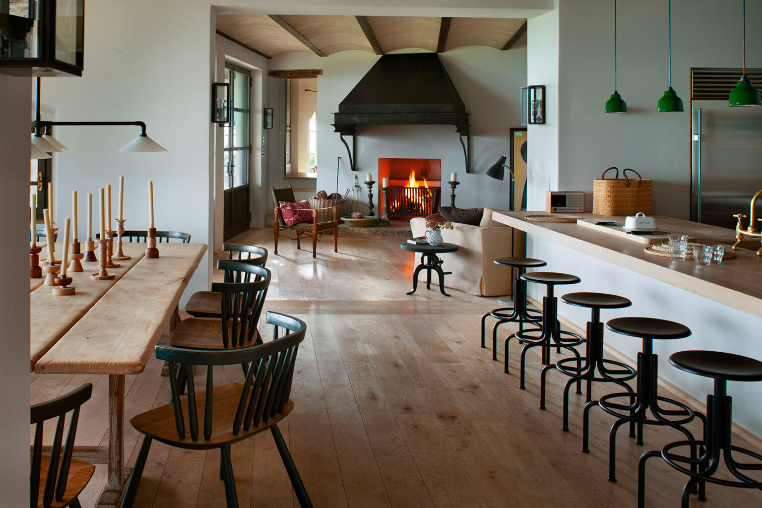Modern rustic interior design featuring a spacious living area with a large fireplace, wooden dining table, and bar seating. The room showcases a blend of natural light, green pendant lights, and a cozy atmosphere, ideal for gatherings and relaxation. | Only on SopranoVillas