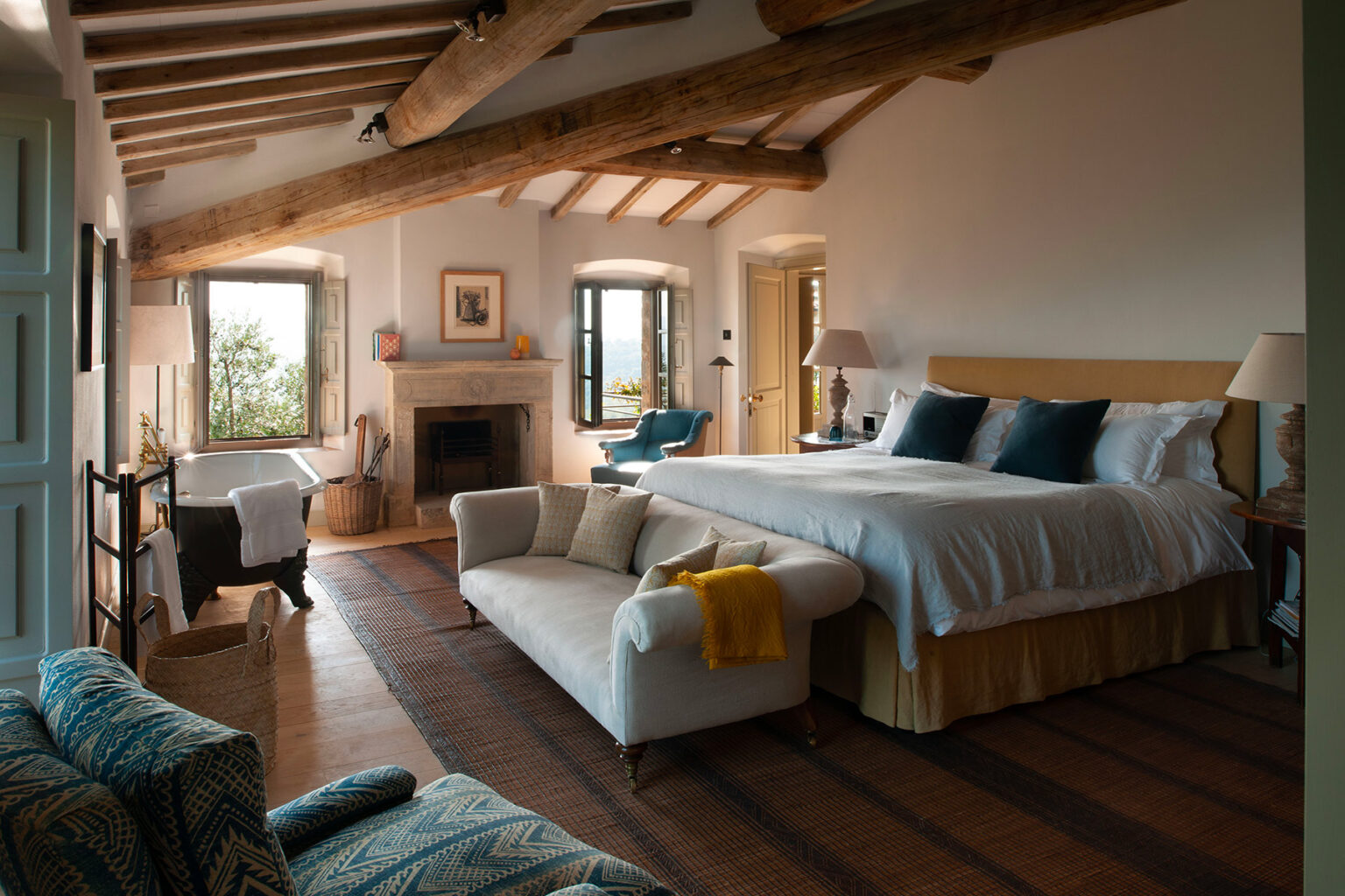 Spacious and inviting bedroom featuring a king-sized bed, cozy seating area with a sofa, natural light from large windows, rustic wooden beams, and a warm color palette, ideal for relaxation and comfort. | Only on SopranoVillas