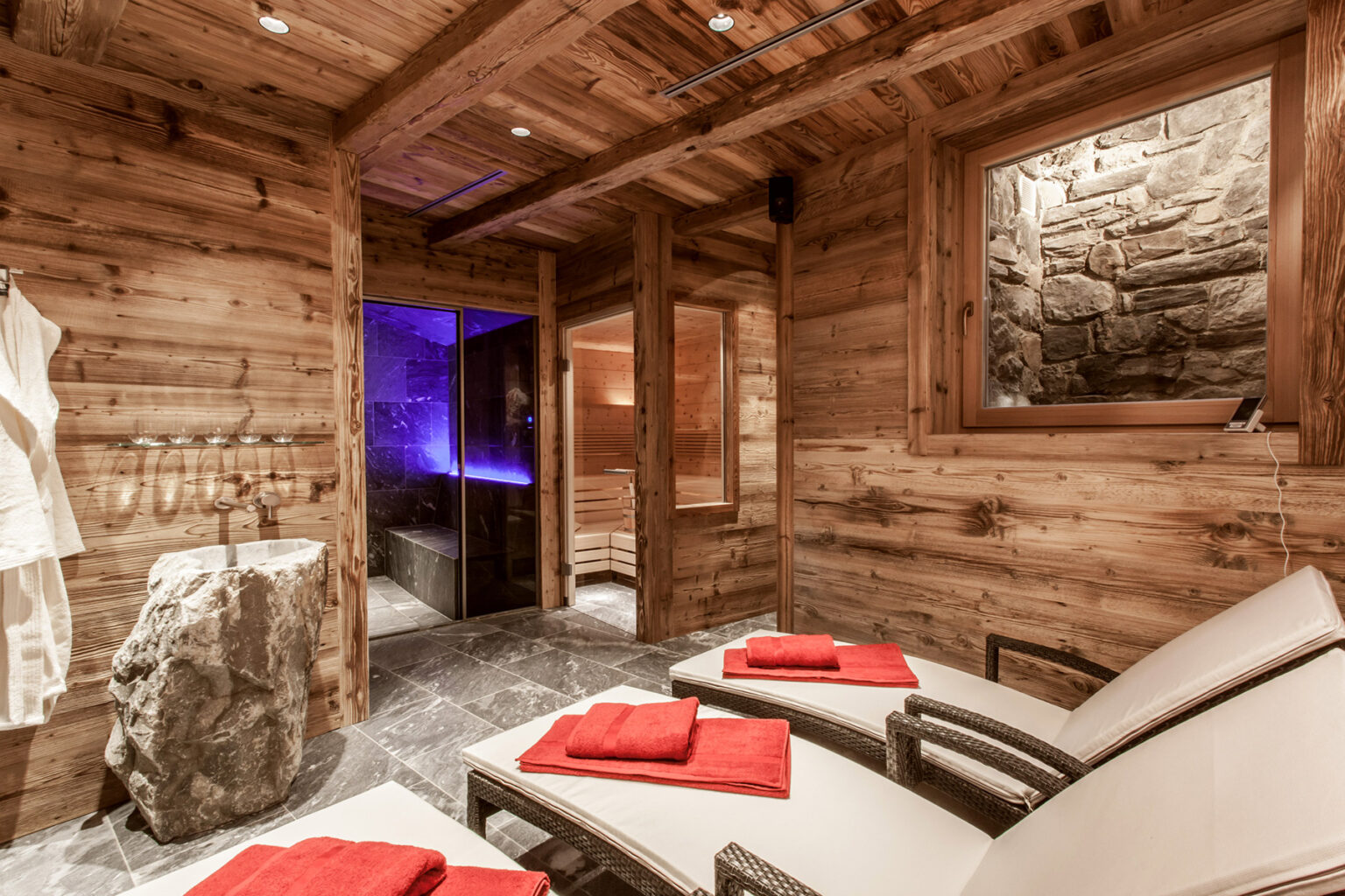Relaxing wooden spa interior with lounge chairs, red towels, and a stone accent wall, featuring a sauna and ambient lighting for a tranquil atmosphere. | Only on SopranoVillas