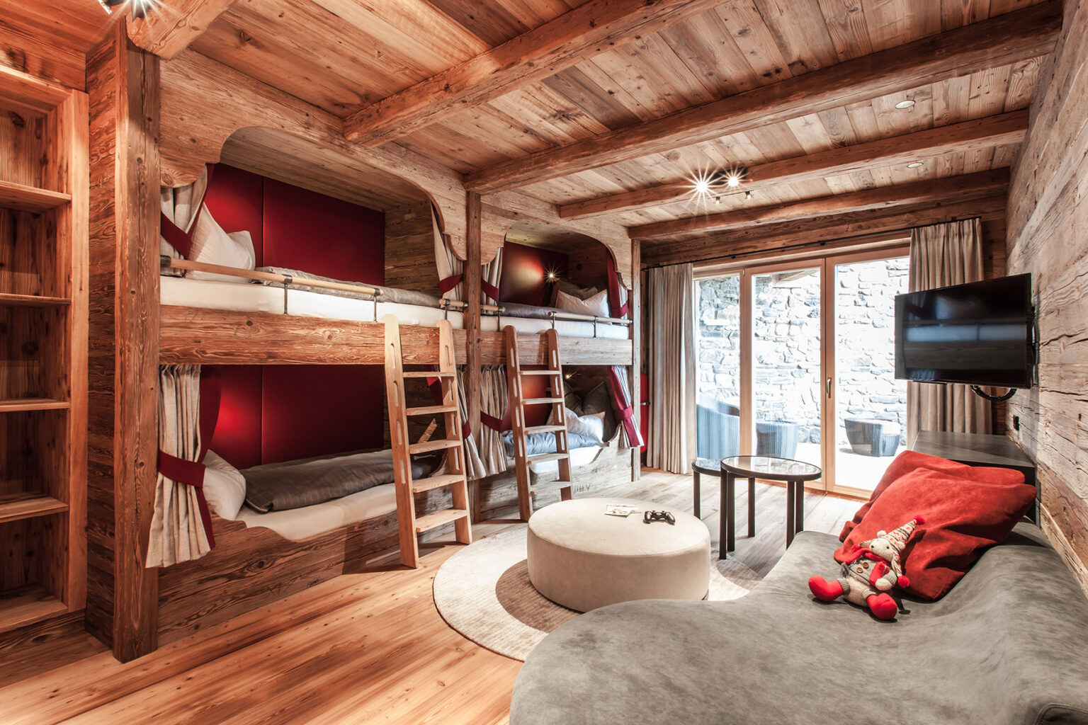 Cozy wooden interior of a modern alpine-style bedroom featuring bunk beds, a round coffee table, and a comfortable lounge area, with large windows providing natural light and outdoor views. | Only on SopranoVillas
