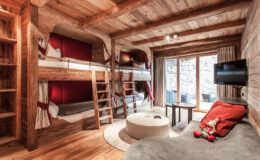 Cozy wooden interior of a modern alpine-style bedroom featuring bunk beds, a round coffee table, and a comfortable lounge area, with large windows providing natural light and outdoor views.