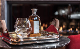 A polished silver tray holds a whiskey bottle and a glass in a cozy setting, with a warm fireplace in the background, creating an inviting atmosphere for relaxation and enjoyment.