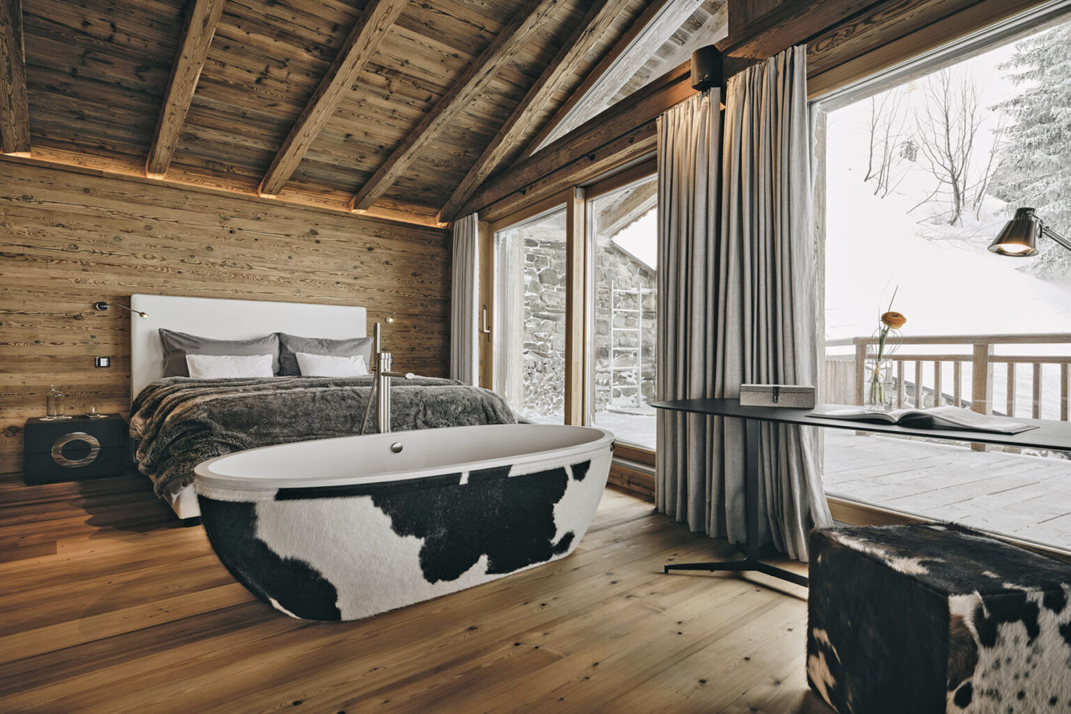 Modern rustic bedroom featuring a stylish cow-print freestanding bathtub, wooden ceiling, and large windows with mountain views. The decor includes a cozy bed with gray linens and a wooden accent wall, creating a warm and inviting atmosphere. | Only on SopranoVillas
