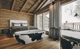 Modern rustic bedroom featuring a stylish cow-print freestanding bathtub, wooden ceiling, and large windows with mountain views. The decor includes a cozy bed with gray linens and a wooden accent wall, creating a warm and inviting atmosphere.