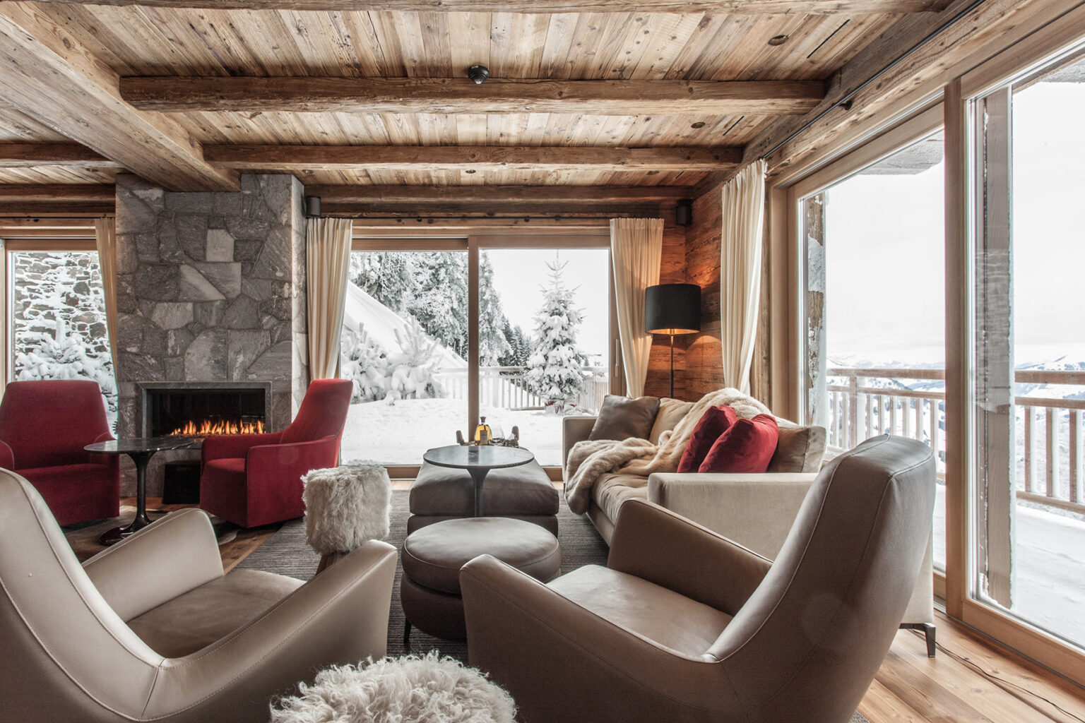 Cozy winter living room with wooden beams, a stone fireplace, and large windows showcasing a snowy landscape. Elegant armchairs and a plush ottoman create a warm and inviting atmosphere. | Only on SopranoVillas