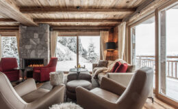 Cozy winter living room with wooden beams, a stone fireplace, and large windows showcasing a snowy landscape. Elegant armchairs and a plush ottoman create a warm and inviting atmosphere.