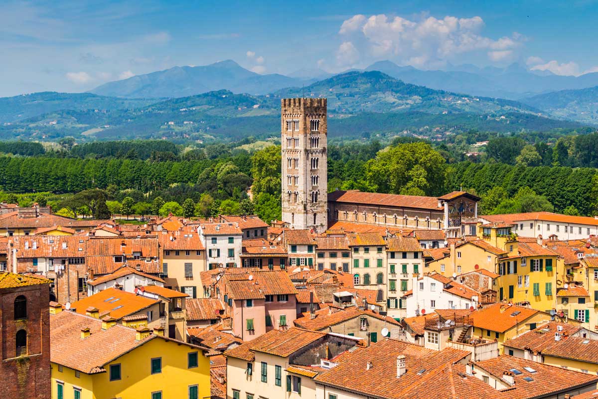 Aerial view of Lucca, Italy, showcasing historic buildings with terracotta roofs, the prominent Guinigi Tower, and lush green mountains in the background under a clear blue sky.