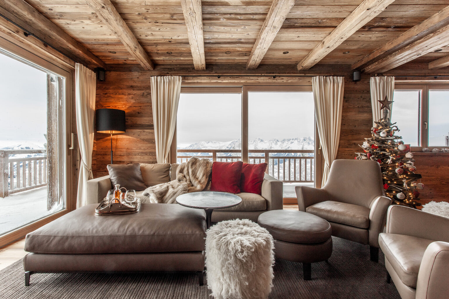 Cozy wooden living room featuring a plush sofa, decorative pillows, and a small Christmas tree, with large windows showcasing a snowy landscape. | Only on SopranoVillas