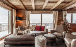 Cozy wooden living room featuring a plush sofa, decorative pillows, and a small Christmas tree, with large windows showcasing a snowy landscape.