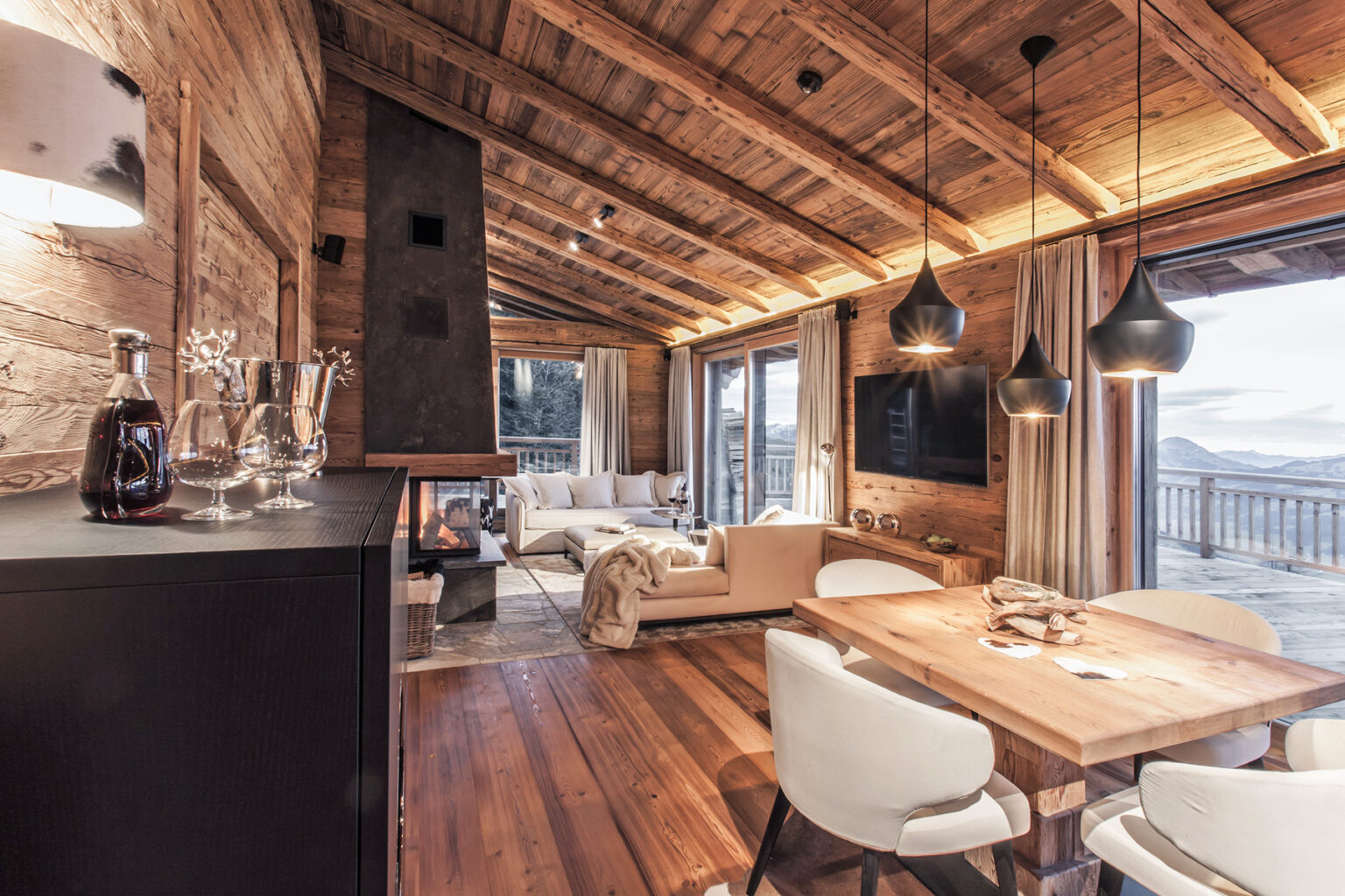Cozy wooden interior of a modern chalet featuring a dining area with a wooden table, stylish chairs, and a living space with a plush sofa. Large windows offer scenic mountain views, while warm lighting and rustic decor create a welcoming atmosphere. | Only on SopranoVillas