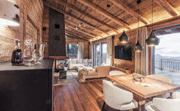 Cozy wooden interior of a modern chalet featuring a dining area with a wooden table, stylish chairs, and a living space with a plush sofa. Large windows offer scenic mountain views, while warm lighting and rustic decor create a welcoming atmosphere.
