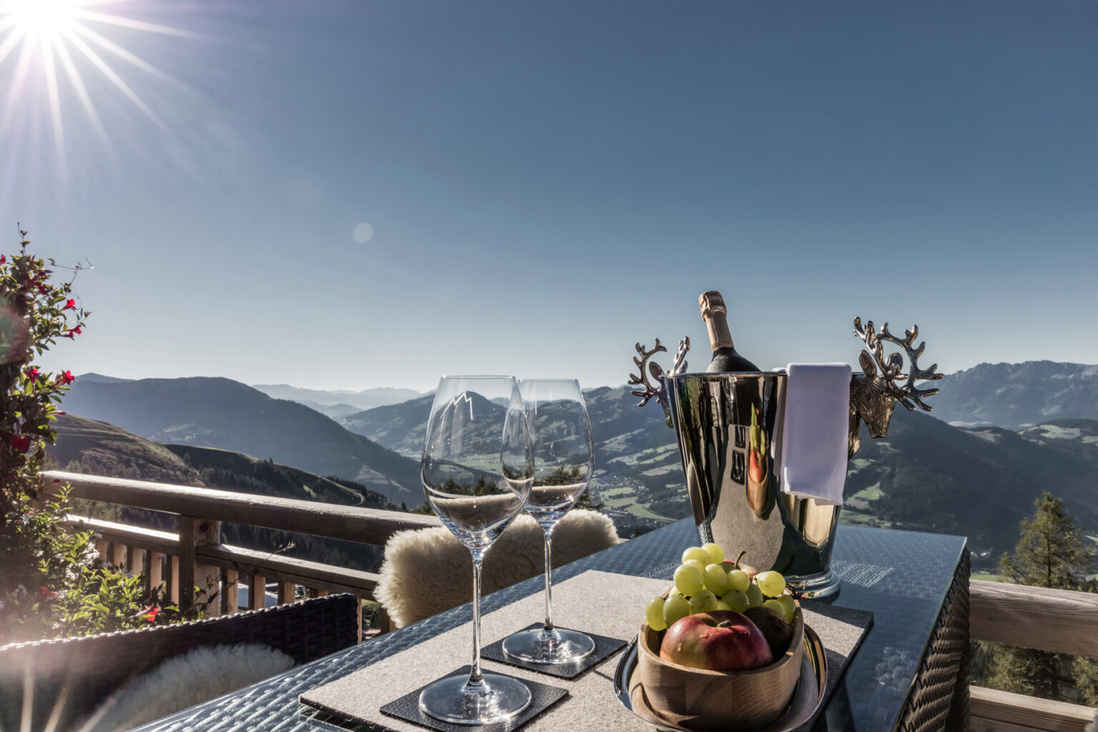Elegant dining setup on a balcony with a stunning mountain view, featuring a bottle of champagne, two glasses, and a platter of fresh fruits. Sunlight bathes the scene, creating a luxurious atmosphere ideal for relaxation and celebration. | Only on SopranoVillas