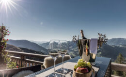 Elegant dining setup on a balcony with a stunning mountain view, featuring a bottle of champagne, two glasses, and a platter of fresh fruits. Sunlight bathes the scene, creating a luxurious atmosphere ideal for relaxation and celebration.